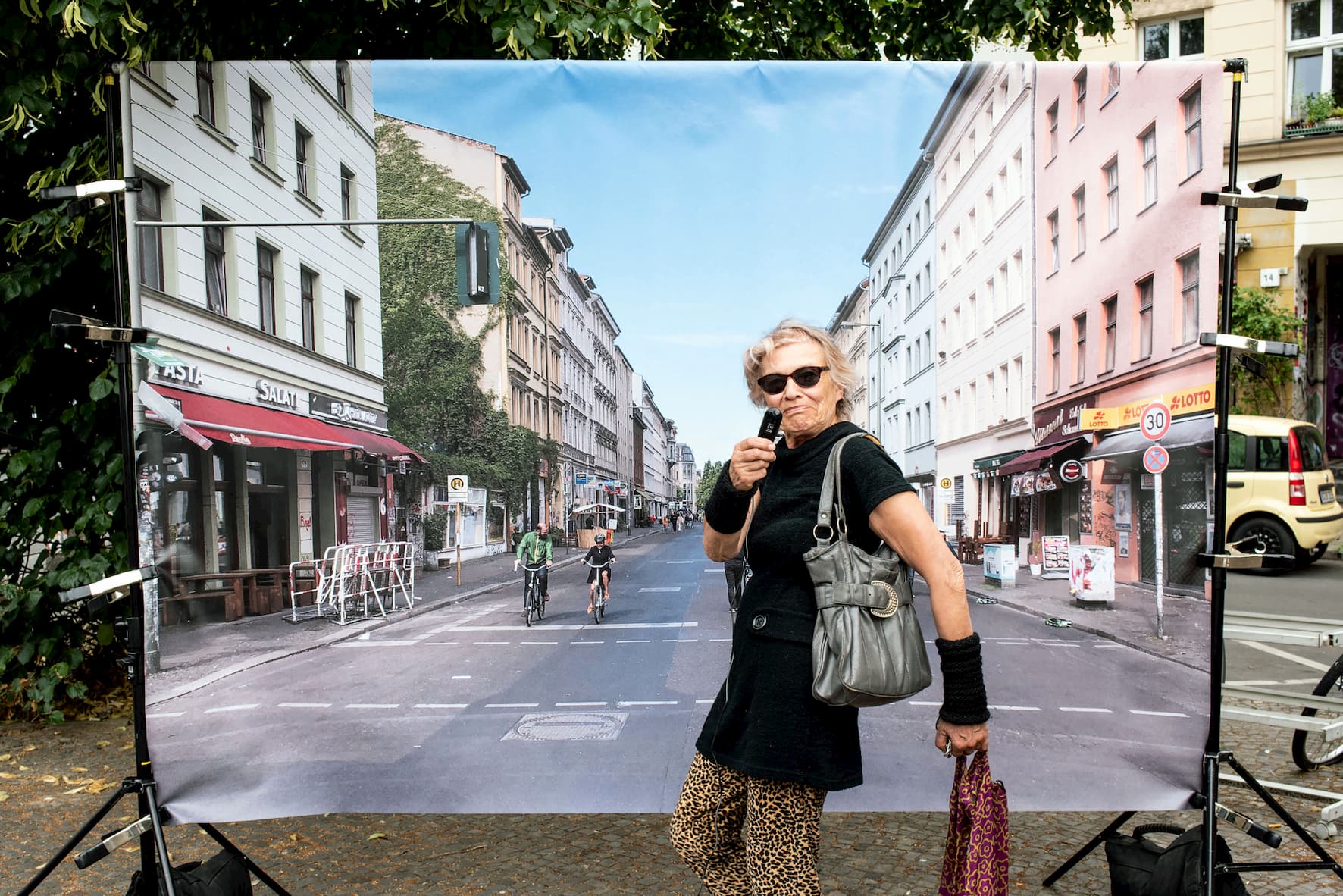 Eine hippe Oma trägt eine Sonnenbrille und posiert vor einer Fotoleinwand. Das Foto ist aus einer Drittperspektive aufgenommen und zeigt eine Straße mit Fahrradfahrern. 