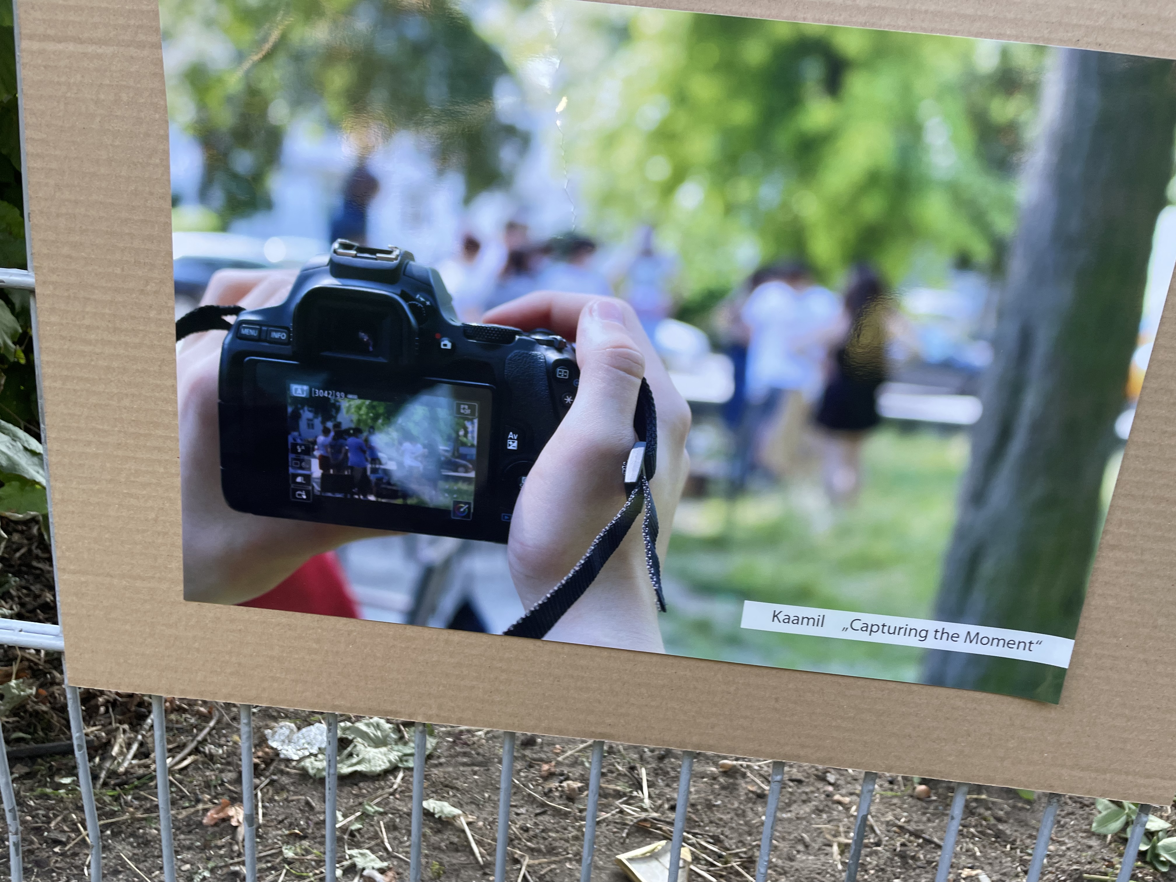 Ein Foto ist ausgedruckt und auf ein Stück Pappe geklebt, das an dem Zaun hängt. Auf dem Foto sieht man eine Spiegelreflexkamera, die die Beteiligten beim Sprayen fotografiert.