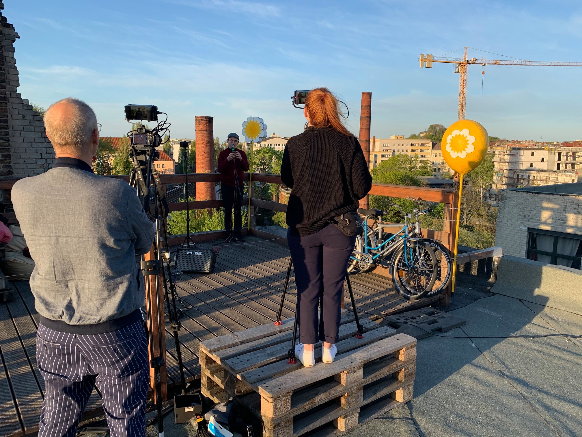 Ein Künstler steht in der Ecke des Daches, davor auf zwei gestapelten Paletten nimmt die Kamerafrau den Livestream auf. Ans Geländer gelehnt stehen Fahrräder.