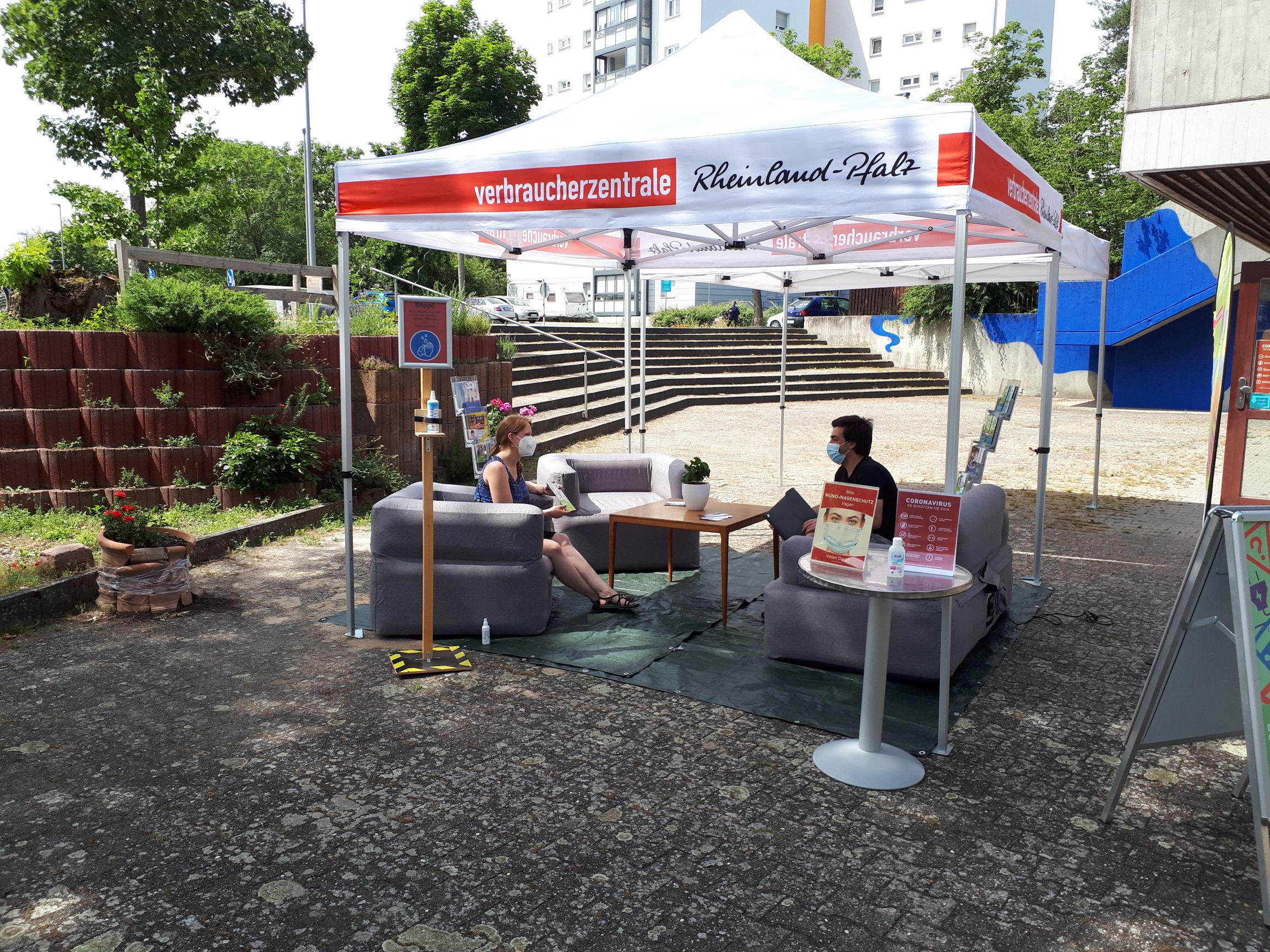 ein weißer Pavillon der VZ Rheinland-Pfalz steht auf einem öffentlichen Platz vor dem Quartiersbüro. Darunter sind drei aufblasbare Sessel um einen Couchtisch angeordnet. Zwei maskierte Erwachsene sitzen sich gegenüber und unterhalten sich.
