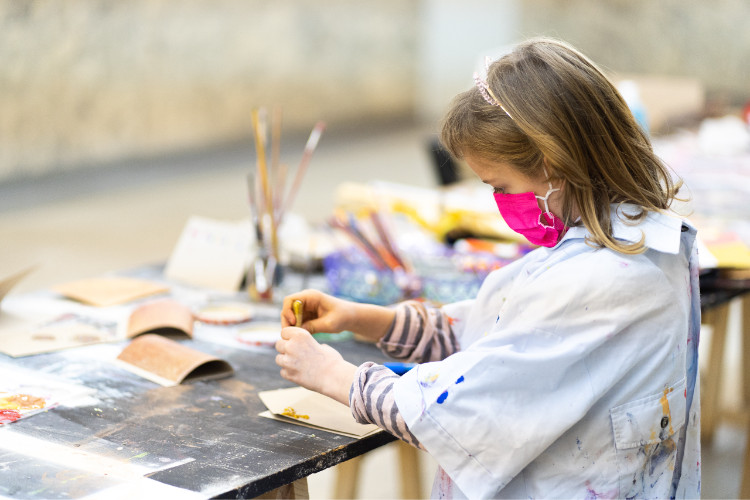 Auf einem Tisch sind Pinsel und diverse andere Bastelutensilien verstreut. Ein Mädchen drückt Farbe aus einer Tube auf ein Stück Papier.