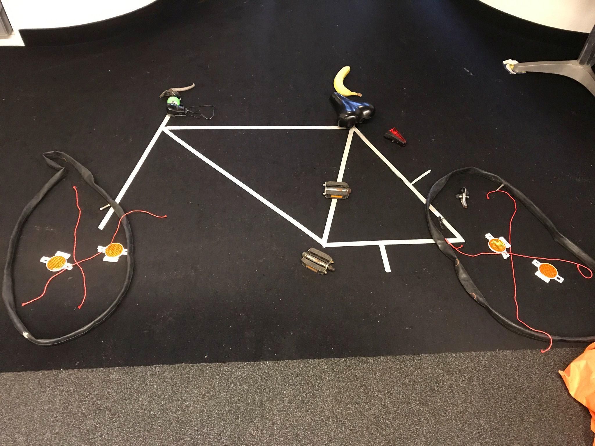 Auf einem schwarzen Untergrund ist mit weißen Klebeband der Rahmen eines Fahrrads aufgeklebt. Die restlichen Teile des Fahrads wurden anders dargestellt: So sind die Speichen rote Fäden, die Pedale sind richtige Fahrradpedale, die Reifen leere Schläuche usw. 