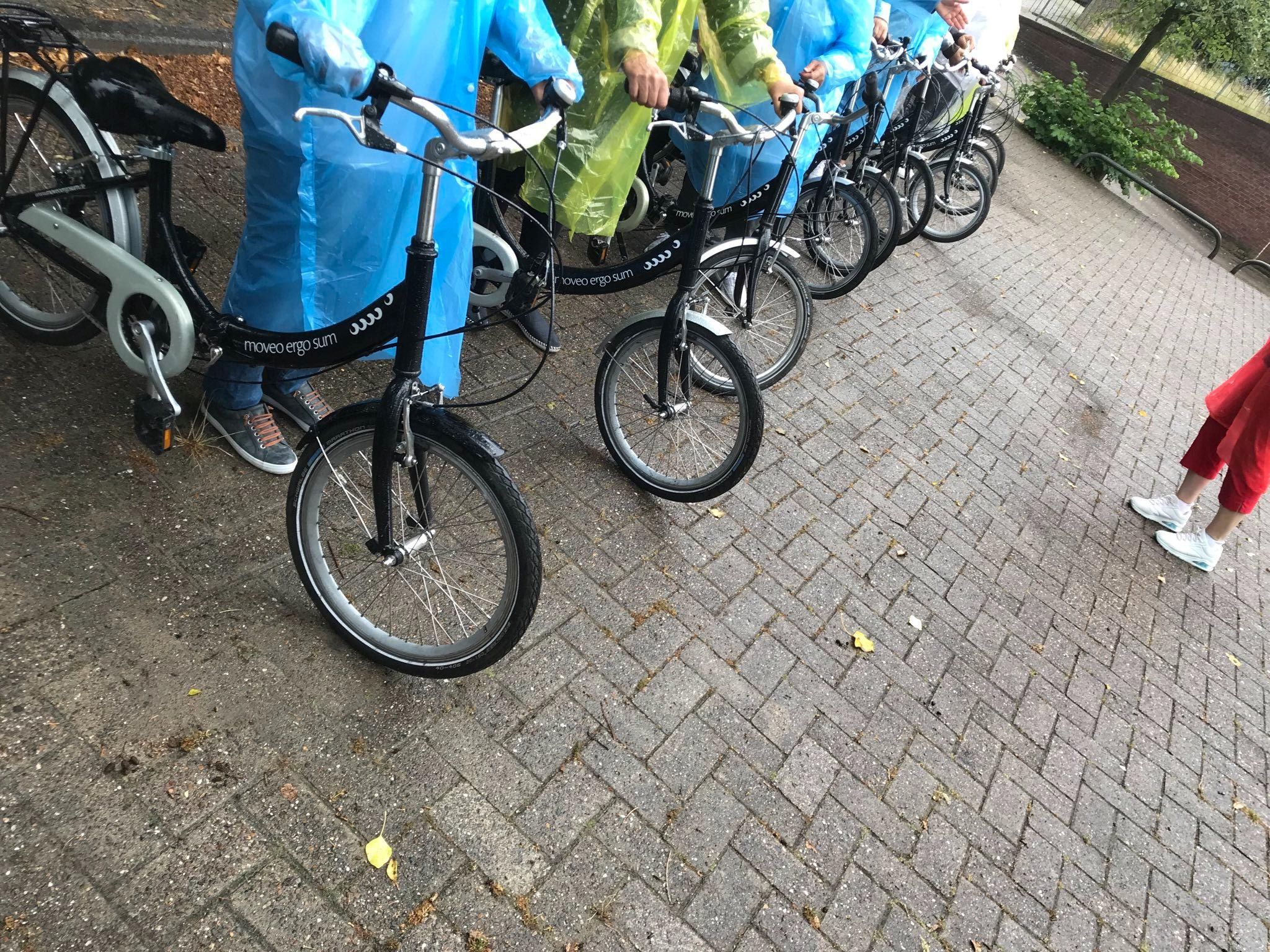Auf einem Platz stehen mehere Personen mit einem Fahrrad nebeneinander. Sie trage alle ein Regenponcho. Alle Personen haben das gleiche Fahrrad. Vor ihnen steht eine Person.
