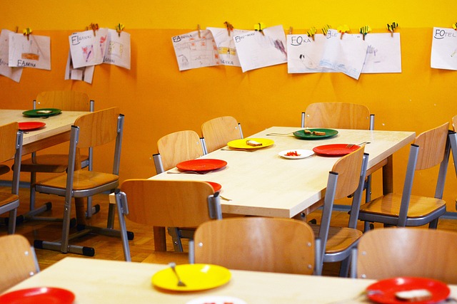 Zu sehen sind mit bunten Tellern gedeckte Tische in einem gelb gestrichenen Raum. Auf manchen Tellern liegt Essen, darunter Brot und Paprika. An der Wand hängen selbst gemalte Bilder, es ist der Speiseraum einer KiTa. 