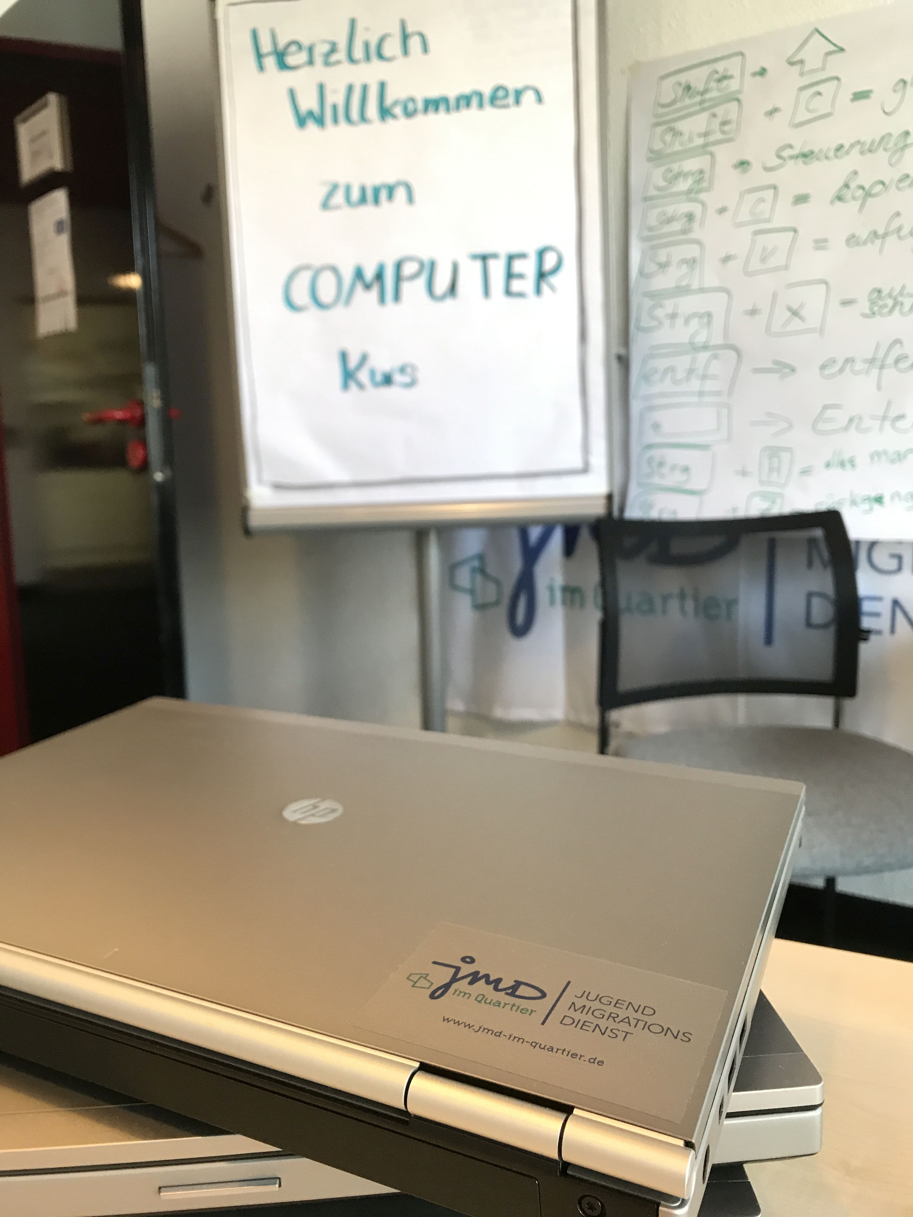 Zwei Laptops liegen übereinander auf einem Tisch. Im Hintergrund steht eine Flipchart aufgestellt mit den Worten: "Herzlich Willkommen zum COMPUTER Kurs".
