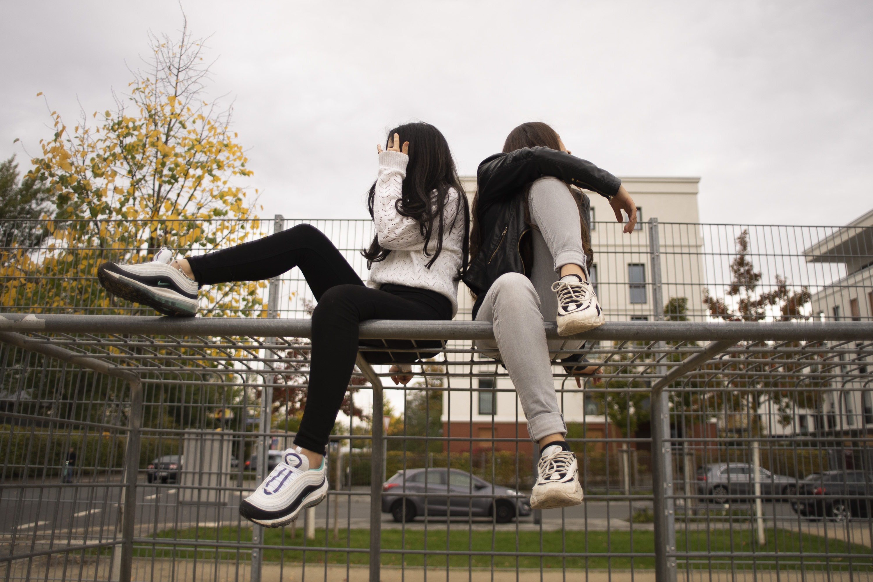 Zwei Mädchen mit langen Haaren sitzen, zur Seite wegguckend und Füße angewinkelt, auf einem Metalltor nebeneinander. 