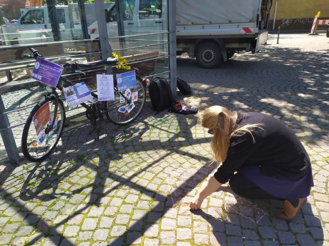 Das Info Fahrrad steht vor einem Sportplatz. Die betreuende Mitarbeiterin knieht davor und malt mit Kreide etwas auf den Boden
