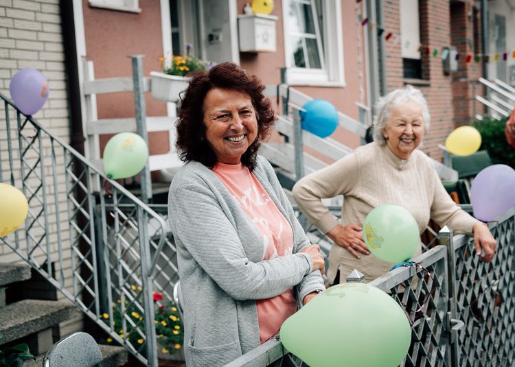 Zwei Omas lachen und stehen hinter einem Zaun in ihrem Vorgarten. Um sie herum sind viele bunte Ballons angebracht.