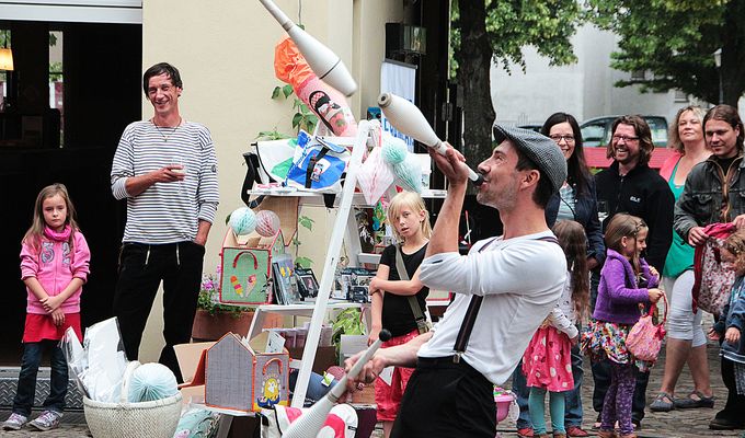 Ein Mann mit Hosenträgern jongliert mit Jonglierkeulen. Um ihn herum stehen Kinder und Erwachsene, die lachen.