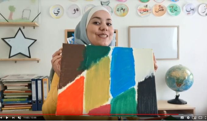 Eine junge Frau mit Kopftuch lächelt und hält ein farbenfrohes Bild vor sich.