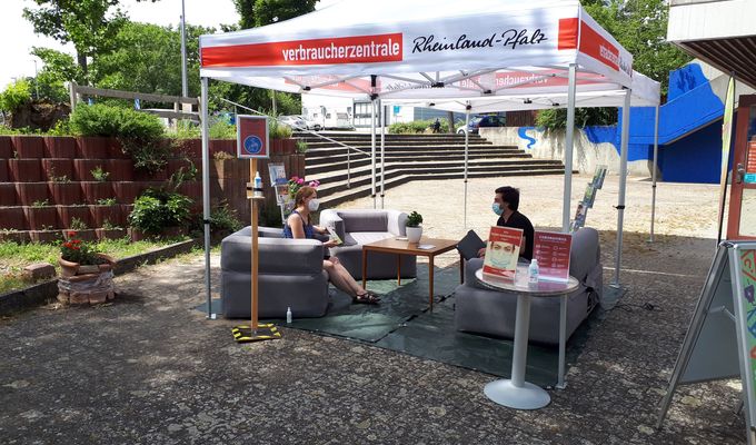ein weißer Pavillon der VZ Rheinland-Pfalz steht auf einem öffentlichen Platz vor dem Quartiersbüro. Darunter sind drei aufblasbare Sessel um einen Couchtisch angeordnet. Zwei maskierte Erwachsene sitzen sich gegenüber und unterhalten sich.