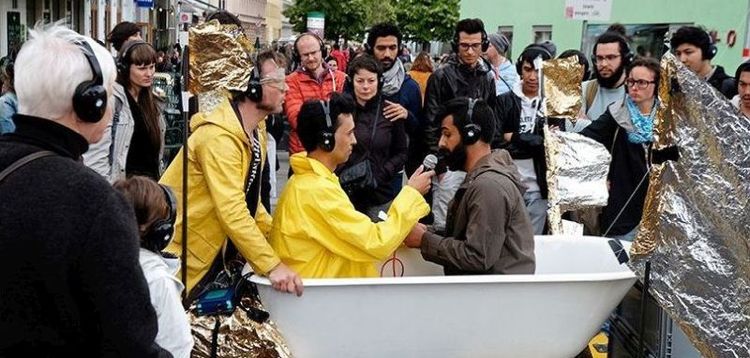 Eine Gruppe von Menschen steht um eine dekorierte Badewanne, in der zwei Personen sitzen. Die eine Person hält ein Mikrofon zu der anderen sprechenden Person. Alle Beteiligten tragen Kopfhörer.
