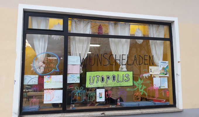 Ladenfenster des Wünscheladens UTOPOLIS. Mit Fingerfarben wurde Geschenke und eine Sprechblase aufs Fenster gemalt. In der Sprechblase steht "deine Meinung zählt".