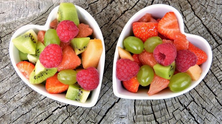 Auf einer Holzoberfläche stehen zwei herzförmige Keramik-Schüsseln die mit einem bunten Obstsalat gefüllt sind. Die Obstsorten sind u.a. Himbeere, Erdbeere, Kiwi, Trauben, Melone und Grapefruit.