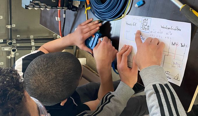 Zwei Jugendliche werkeln stehend an einem technischen Gerät herum. Einer steckt ein Kabel ins Gerät, der andere zeigt auf eine selbst erstellte, technische Skizze. 