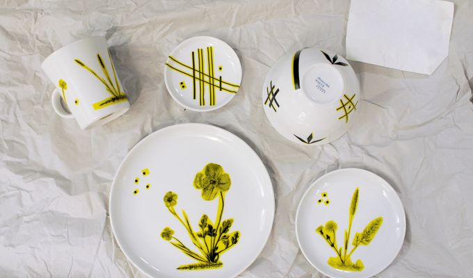 Drei bemalte Teller und zwei Tassen liegen auf einem weißen Hintergrund. Das Geschirr ist mit gelb-grünlichen Mustern und Pflanzen bemalt.