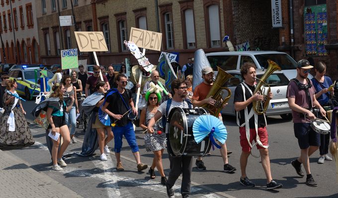 Eine große Gruppe junger Personen laufen verkleidet eine Straße entlang. Einige von ihnen tragen Schilder oder spielen Instrumente. 