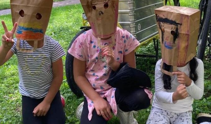Drei Kinder sitzen auf dem Boden und haben eine Papiertüte auf dem Kopf, auf welcher ein selbstgemaltes Gesicht drauf ist. Hinter den Kindern steht ein Lastenrad. 