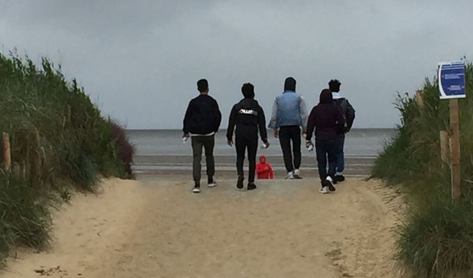 Fünf Männer sind von hinten zu sehen, wie sie einen breiten Sandweg entlang auf das Meer zugehen. Rechts und links vom Weg sind Dünen mit Strandhafer-Bewuchs. Der Himmel ist grau. 