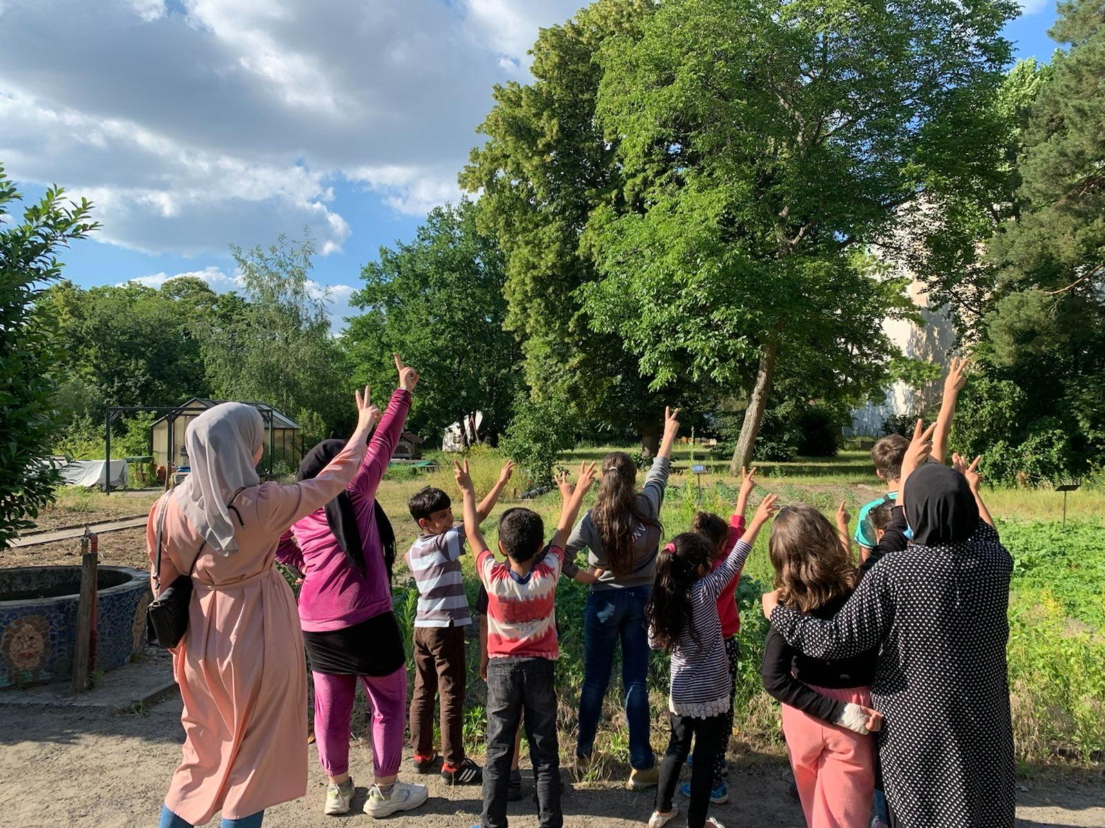 Kinder und Erwachsene stehen vor einem Garten mit Beeten und einem Gewächshaus. Alle Personen strecken ihre Hand nach oben und zeigen das Peace-Zeichen.