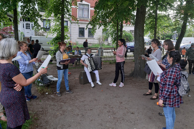In einem Park stehen Menschen in einem Halbkreis. Ein Mann spielt auf einem Akkordeon. Links und rechts von ihm stehen Menschen mit einem Liedblatt in der Hand und singen mit.