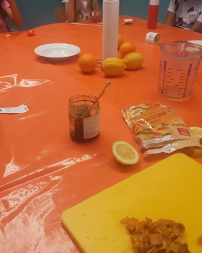 Auf einem Tisch mit knallorangener Plastiktischdecke liegen Zitronen, Orangen, Mangostreifen, ein Messbecher, Küchenrolle, ein Teller sowie ein Schneidebrett mit Obst. Am Bildrand ist gerade noch zu erkennen, dass um den Tisch Kinder sitzen